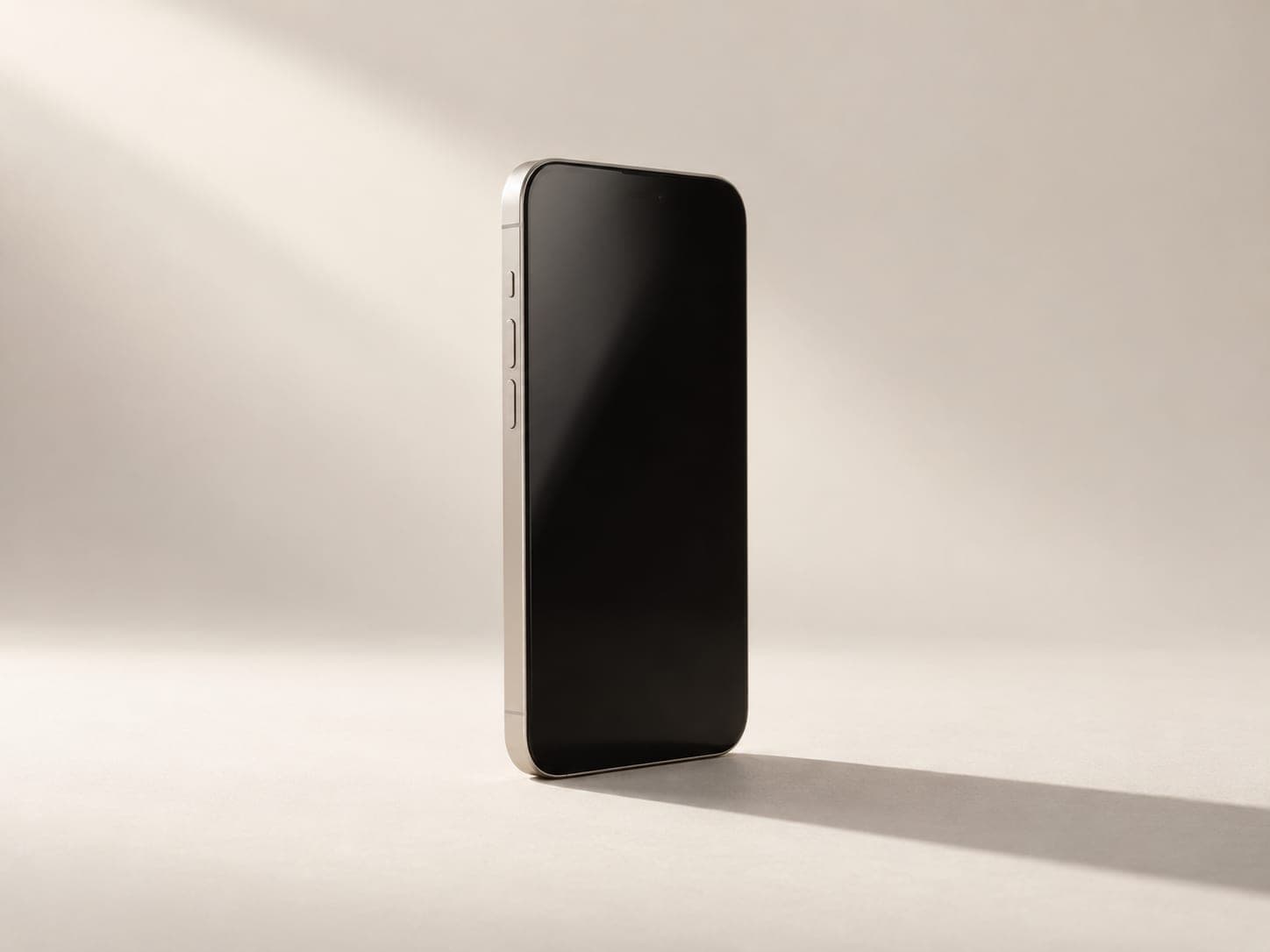 Un téléphone debout, écran éteint, sur un fond ivoire traversé par la lumière d’une fenêtre.